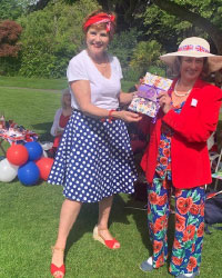 A woman in a spotty skirt, white top and red head scarf smiling with a woman in a red blazer 