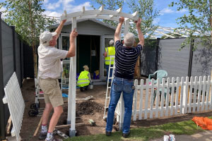 Two men building a white wooden arch. Behind them is an empty plot, in which two people in yellow high vis jackets are standing looking at a shed.