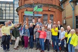 A large group of older adults, many wearing blue and yellow, are standing in front of a sign post pointing to it. They are standing on the streets of London in front of an old building.