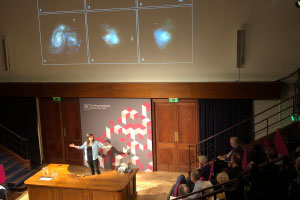A woman with outstretched arms stands in a crowded auditorium, in front of a screen that shows galaxies