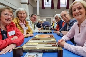 A group of older adults are sat around a table, smiling at the camera. The table has a game on it.