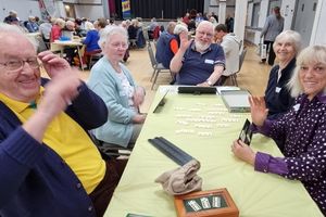 Five older adults sitting around a table that has a game of dominos on it. They are looking at the camera and waving
