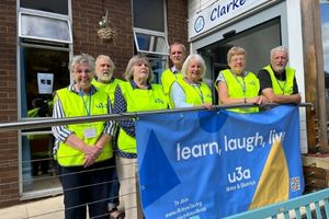 Seven older adults wearing high vis vests that say u3a are standing on a ramp into a building. In front of the rap is a big banner that says 'learn, laugh, live' above the 'u3a Ilkley & District' logo