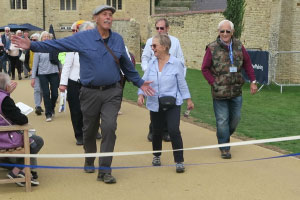 A group of older adults are walking on a path outside with a ribbon going across it. The man at the front has his arms opened wide and is smiling. Next to him, a woman is looking to the left and laughing.