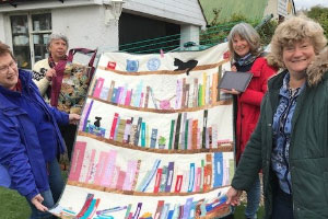 Four women holding up a quilt with bookshelves and a cat on.
