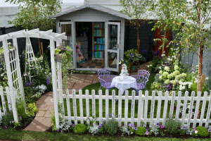 A white picket fence and a white arch lead to a show garden with green lawn, purple and white flowers. On the lawn is a white table with a two tiered cake - on the top tier it says "40" and on the bottom tier "u3a." Around the table are two purple chairs. A path leads to a shed which has open doors. Inside the shed, there are shelves, a book case, a model boat and a quilt.