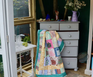 A set of drawers is open, with a patchwork quilt coming out. On top of the drawers are several items, including a pot of paintbrushes and a purple jug with white flowers coming out. On the floor there are two tennis rackets. On the other side of the drawer is a white table with empty glasses and grapes. Beneath this table are a pair of walking boots.