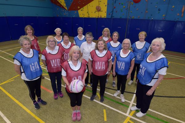 Women standing inside on a netball pitch; some are wearing blue bibs, some are wearing red bibs. The woman in the centre is holding a ball. They are all facing towards the camera and smiling.