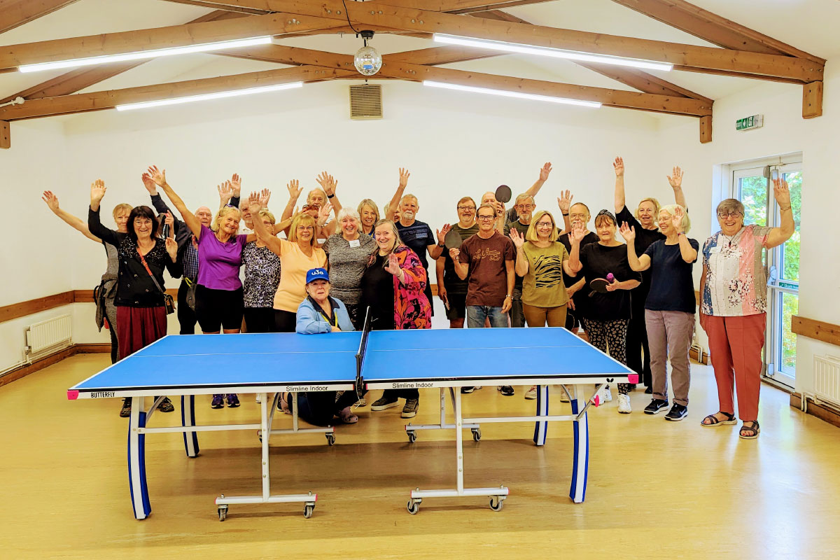 A large group of people standing in a hall, behind a table tennis table, with their hands outstretched in the air