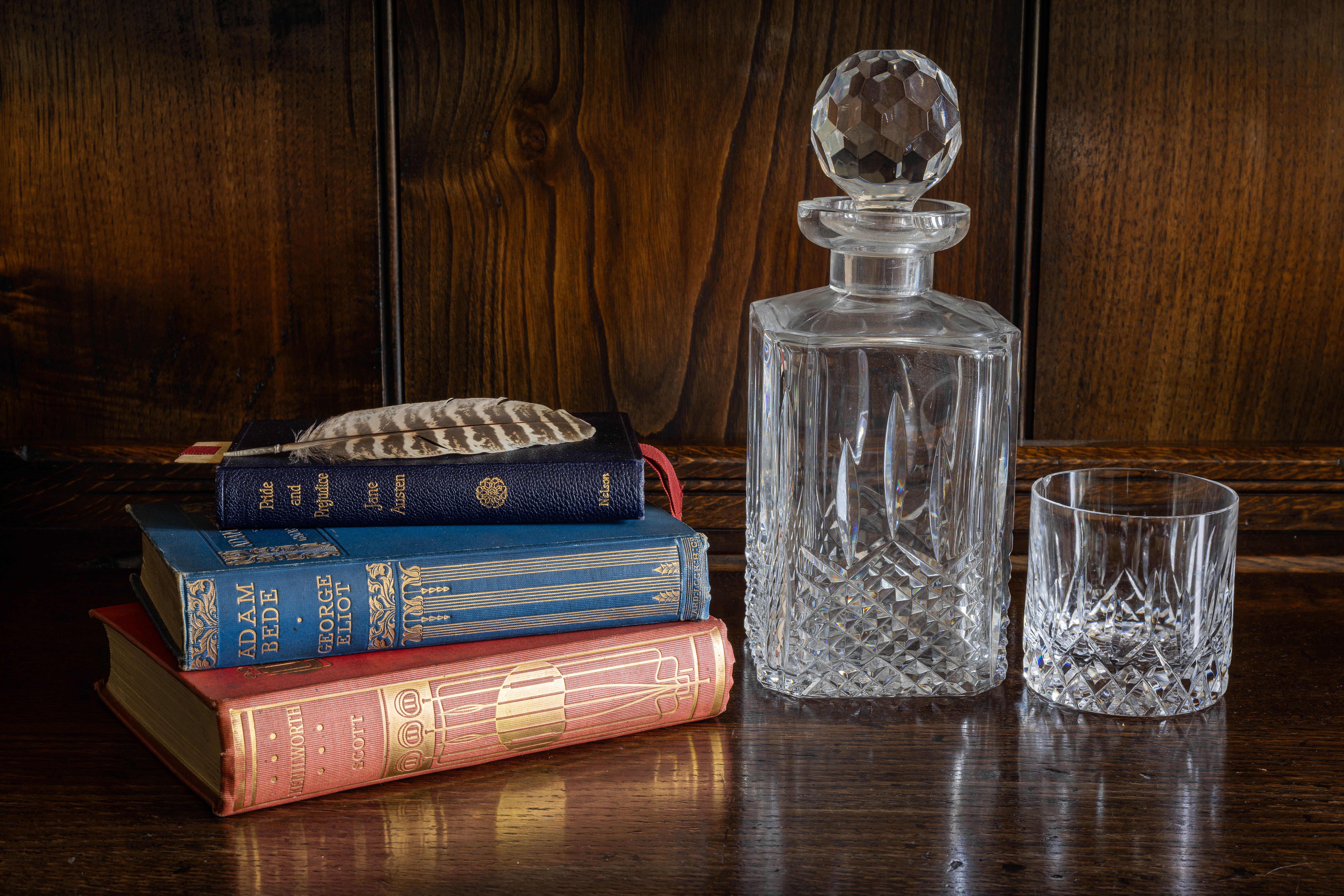 3 old fashioned books piled on the left, on a dark wooden table. A crystal decanter and glass are on the right 