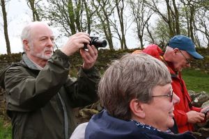 Two men and a woman standing outside in the countryside - they are standing next to an old stone wall. They are looking at things in the distance and one man is holding up a camera to take a photo; the other looks like he is looking down at a photo he has just taken on his camera.