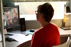 A woman in a red cardigan sits in a home office in front of a computer. The computer is showing a Zoom call with nine faces, who look like they're involved in a discussion.