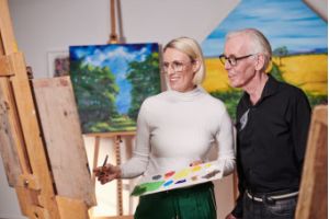 A woman wearing glasses holding a paintbrush in front of a canvas and an older man looking at the canvas