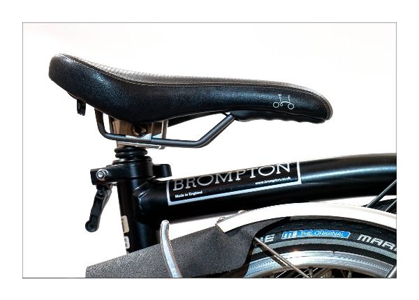 A bicycle seat, with part of the bicycle bar and wheel showing, the Brompton bike logo is also visible. 