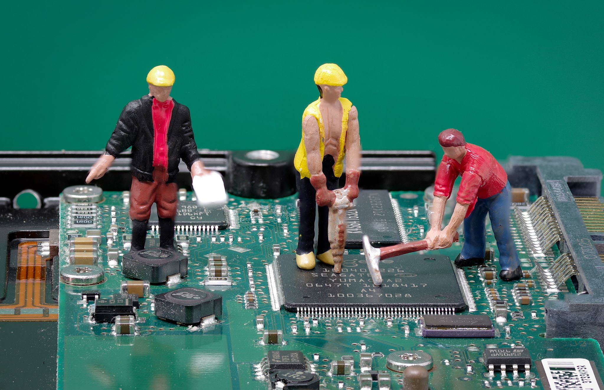 A green and black circuit board with minitature figures of workmen on it