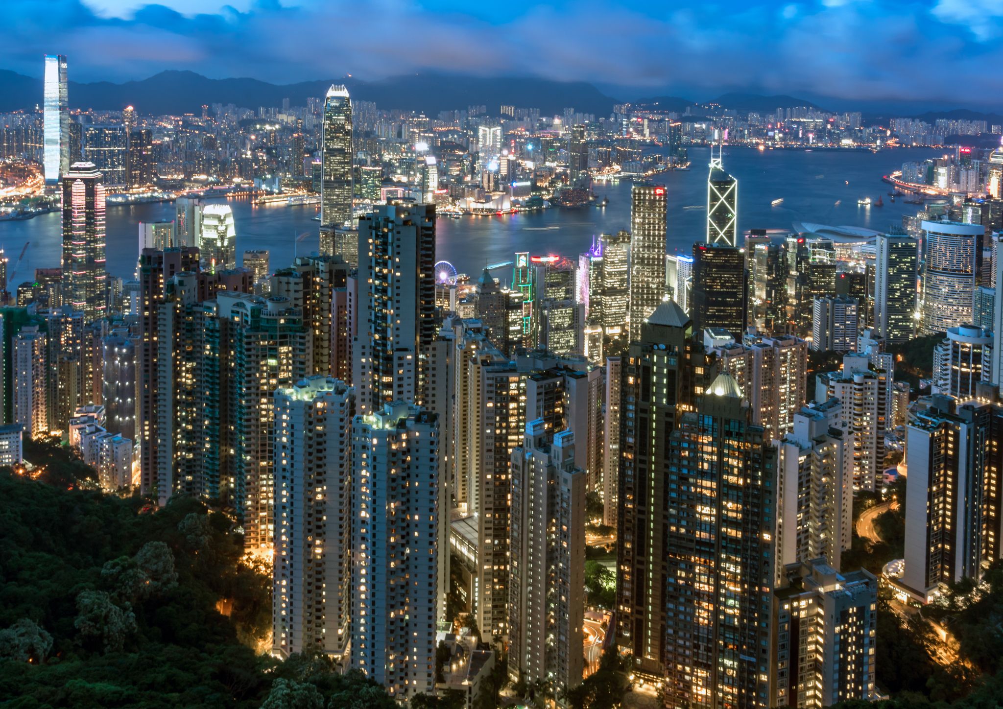 very tall buildings close together in a city, it's getting dark and the windows of the buildings are lit up yellow. Water in the background. 