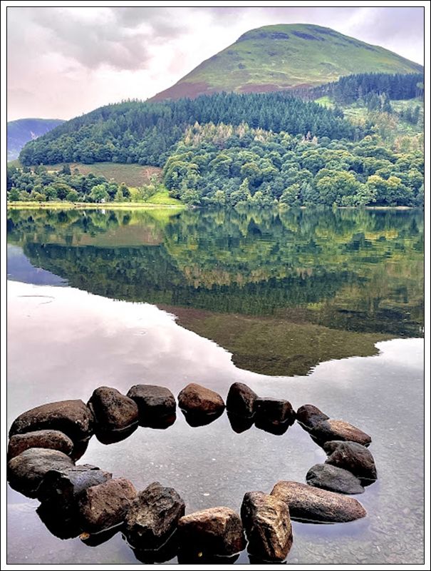 A circle of rocks in front of a loch with a wooded hill in the background