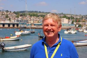 A man with blonde hair and wearing a bright blue top is standing in front of a harbour on a beautiful day.