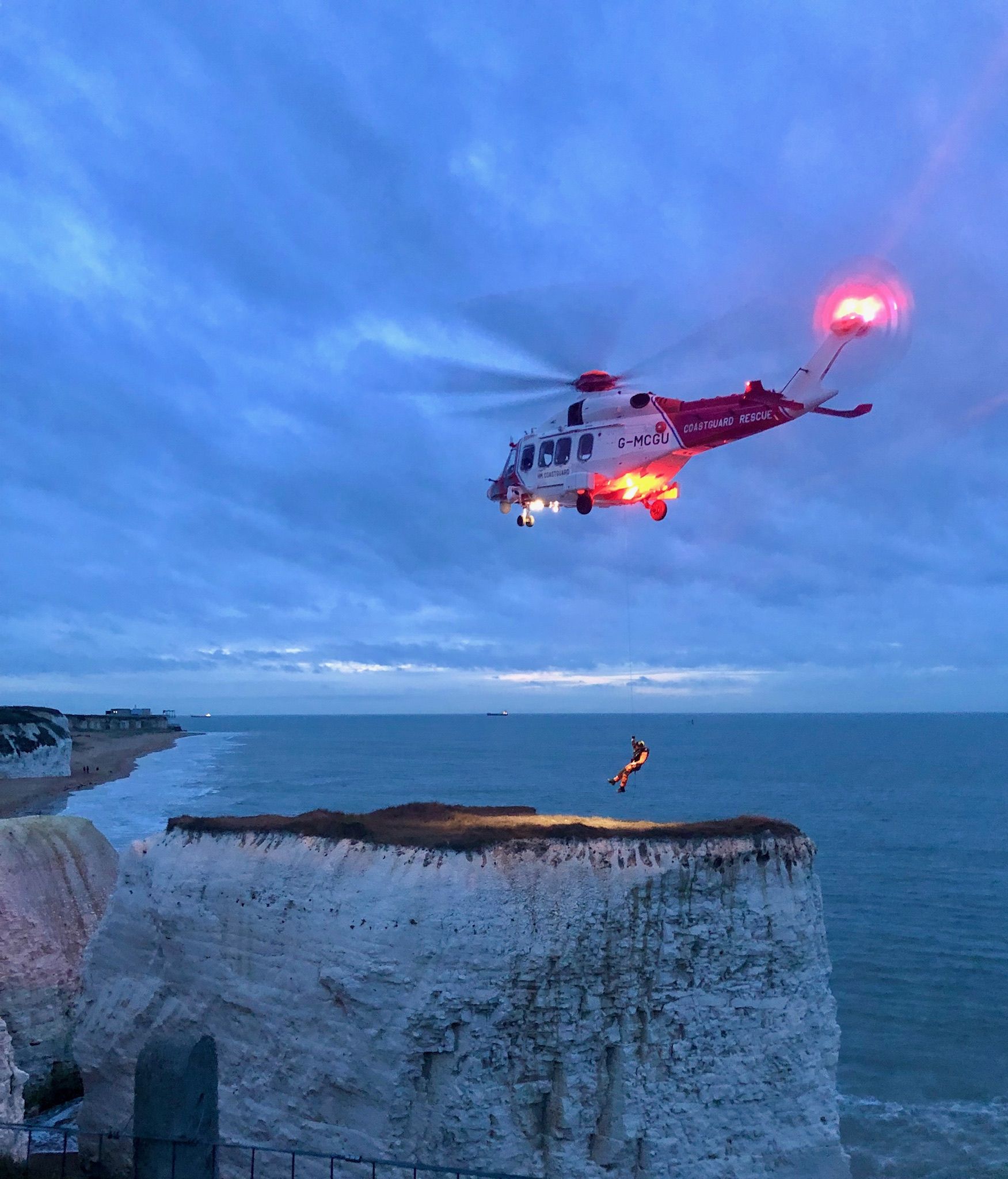 A photo of a cliff, with a helicopter hovering over it. It is dusk. A person is coming down from the plane to land on the cliff. 