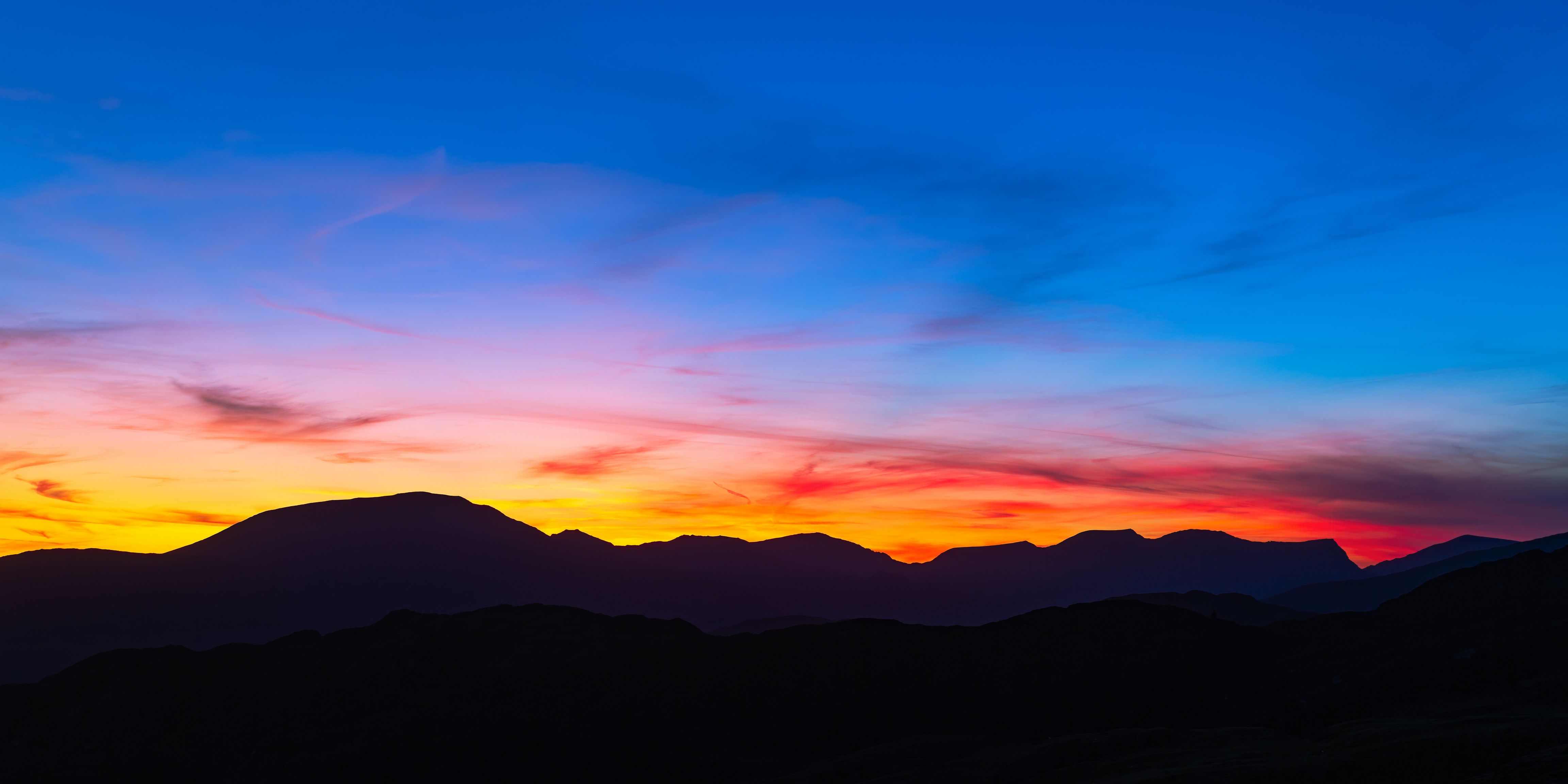 blue sky at the top, with yellow sun setting behind a foregroud of silhouetted mountains 
