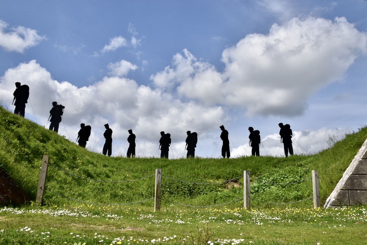 Looking up at a ridge with grass covering it, the shapes of sculptures of men are on the skyline. blue sky in background with white fluffy clouds. 